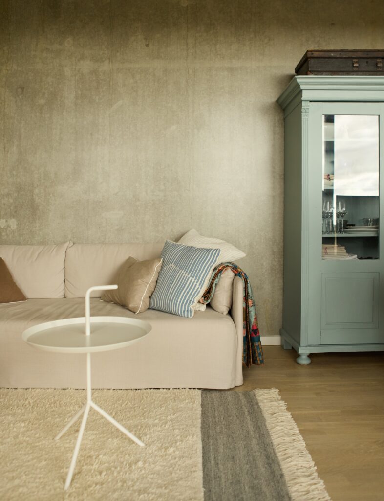 Stue med rå betonvæg, sofa i lys farve med bløde puder og vitrineskab i blå farve
