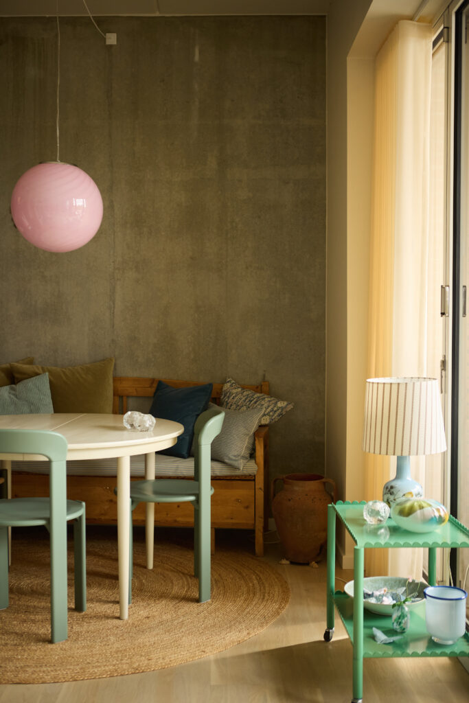 Moderne nybyg-interiør uden vindueskarm med gulv-til-loft vindue. En blød og personlig indretning med bænk langs væggen, rundt spisebord, grønne stole, taktile tekstiler, planter og belysning placeret på møbler og flader i stedet for vindueskarm.